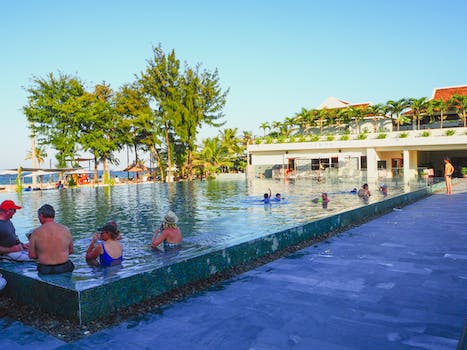 Back view of unrecognizable tourists in swimwear relaxing in outdoor pool on territory of tropical hotel near seashore against cloudless blue sky
