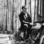 Black and white pensive man in sunglasses sitting on stylish motorcycle in sunny forest