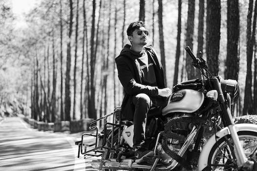 Black and white pensive man in sunglasses sitting on stylish motorcycle in sunny forest
