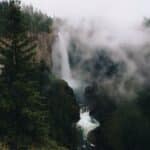 Waterfall in mountainous terrain with evergreen forest against cloudy sky