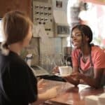 Smiling African American waitress wearing casual t shirt and apron standing at counter in cozy coffee shop with female customer while serving hot drink and looking at each other