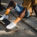 Crop anonymous African American male skater in t shirt and denim shorts sitting on sidewalk with feet on skateboard