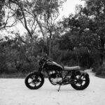 Black and white of metal motorcycle parked on sandy road near forest with deciduous trees and lush bushes
