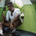 Sad African American male traveler resting in tent and browsing Internet on cellphone while spending journey in nature