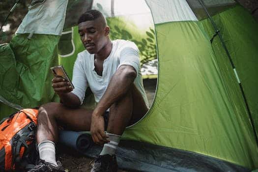 Sad African American male traveler resting in tent and browsing Internet on cellphone while spending journey in nature