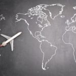 Top view of miniature airplane placed on over gray world map with crop hand of anonymous person indicating direction representing travel concept