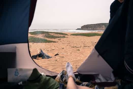 Unrecognizable tourist resting in tent with opened door in nature on sandy seashore with rippling sea and rocky cliff in distance
