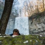 Young male hiker hiding behind boulder near waterfall in forest