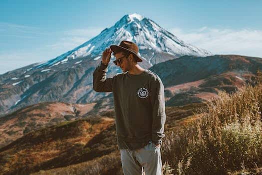 Young stylish man in hat and modern sunglasses standing on hill with hand in pocket against high snowy mountain