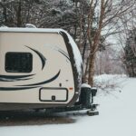 Camper trailer parked in coniferous and deciduous forest on road trip in snowfall in gloomy weather in winter in daytime